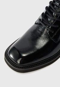 Zapato de cuero negro con un acabado brillante, puntera redonda y cordones negros. La suela tiene un borde texturizado con detalles de costura decorativa.