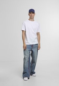 Młody mężczyzna stoi w niebieskiej czapce, białej koszulce, szerokich niebieskich dżinsach z czerwonymi przeszyciami i białych trampkach na jednolitym tle.