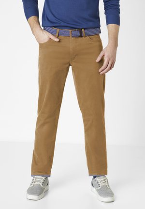 Man wearing camel-colored pants, blue patterned belt, navy long-sleeve shirt, and gray sneakers, standing with one hand in pocket.