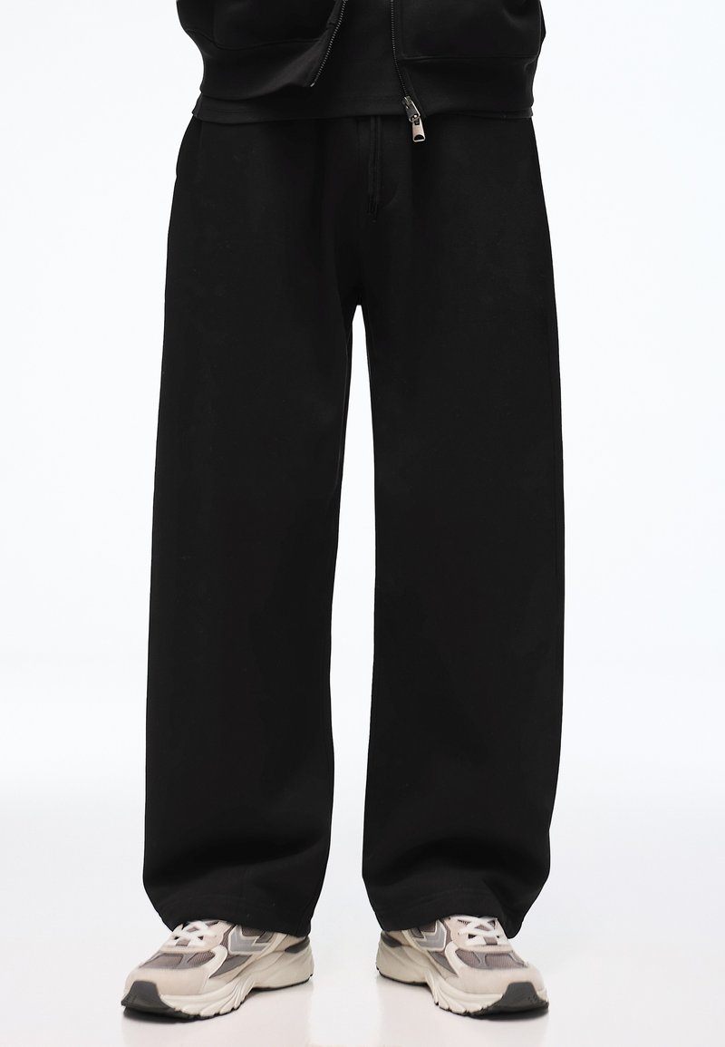 Personne portant un pantalon noir ample et des baskets blanches et grises, debout devant un fond blanc.