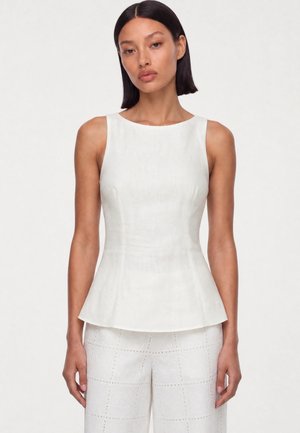 Femme aux cheveux lisses et foncés portant un top péplum blanc sans manches et un pantalon blanc à motifs, debout devant un fond uni.