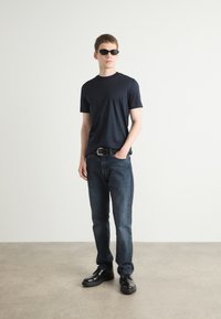 Maglietta navy, jeans blu scuro, cintura nera, scarpe nere. Il modello è in piedi contro uno sfondo neutro, indossando occhiali da sole e con le mani in tasca.