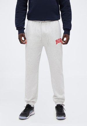 Homme portant un pantalon de jogging gris clair avec des lettres rouges sur la cuisse gauche et des chaussures de sport noires, debout, les mains détendues le long du corps.