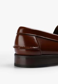 Mocasines de cuero marrón con acabado brillante, tacón redondeado y suela negra en contraste. Presenta costuras decorativas a lo largo de la costura superior.