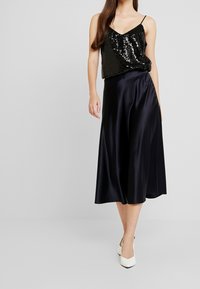 Schwarzes Paillettenoberteil mit dünnen Trägern, kombiniert mit einem fließenden schwarzen Satin-Midirock, das über eine glatte Textur und einen subtilen Glanz verfügt. Weiße High Heels runden den Look ab.