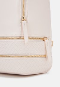 Mochila de cuero rosa con un fondo texturizado trenzado. Cuenta con dos cremalleras doradas y tiradores de cremallera de cuero plano. Superficie suave con acentos mínimos.