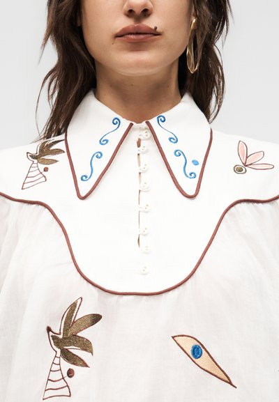 Γυναίκα που φοράει λευκό πουκάμισο με μυτερό γιακά, μπλε σχέδια στροβίλων με κέντημα, καφέ περίγραμμα, μπροστινό μέρος με κουμπιά και αφηρημένα κεντημένα λουλουδένια σχέδια.