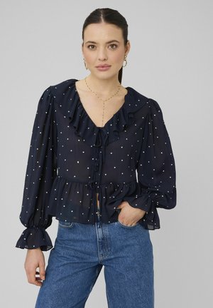 Femme portant un chemisier bleu marine transparent à pois blancs, col à volants, et un jean bleu, posant avec une main dans la poche devant un fond gris.