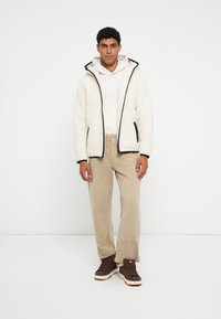 Valkoinen fleece-takki mustalla reunuksella, beige huppari alla, vaaleanbeige housut ja tummanruskeat tennarit. Yksinkertainen, rento muotoilu.