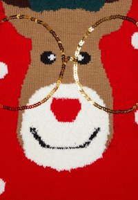 Virkat renhjortsansikte med röd näsa och vit snut på röd bakgrund, prytt med runda guldglitterutramade glasögon.