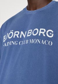 Modré bavlněné tričko s velkým bílým, opotřebovaným potiskem, který zní "BJÖRN BORG TRAINING CLUB MONACO." Kružkový výstřih.