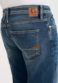 Achteraanzicht van blauwe denim jeans met meerdere gestikte zakken en een bruine leren merkpatch met het label "Le Temps des Cerises."