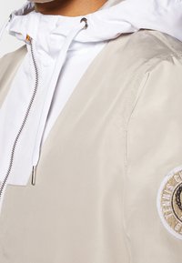 Gros plan d'une personne portant une veste beige par-dessus un sweat à capuche blanc zippé avec un cordon et un patch circulaire beige et blanc sur la manche de la veste.