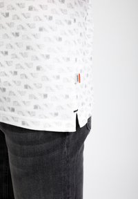 Chemise blanche texturée avec motif géométrique gris et petite fente latérale, associée à un jean en denim noir sur fond blanc.