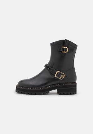Bottine en cuir noir avec semelle épaisse, boucles dorées et sangle tressée décorative sur fond uni.