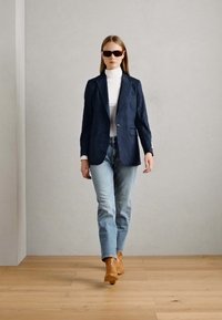 Navy pinstripe blazer over a white turtleneck, paired with light blue jeans and tan ankle boots. Model wears dark sunglasses.