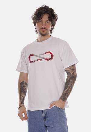 Uomo con capelli ricci che indossa una maglietta bianca con un motivo a simbolo dell'infinito rosso e grigio, jeans blu e tatuaggi dettagliati sulle braccia.