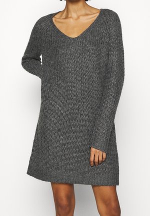 Vrouw draagt een lange, grijze, textuurrijke gebreide trui-jurk met een V-hals en lange mouwen tegen een effen witte achtergrond.