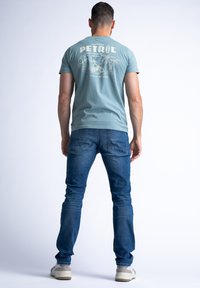 Lichtblauwe katoenen T-shirt met witte grafische print op de achterkant, in combinatie met donkerblauwe jeans en lichte sneakers, gezien vanaf de achterkant.