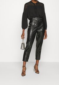 Blouse noire avec détails en dentelle et col noué, associée à un pantalon taille haute en cuir synthétique et des talons noirs à brides. Tenant un sac argenté.