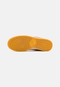 Könnyű, világosbarna felsőrésszel rendelkező sneaker, amelynek sárga gumitalpa körkörös és rácsos mintázattal díszített, és a saroknál egy domború Nike logó található.
