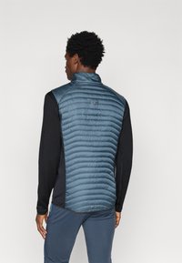 Μπλε αδιάβροχο γιλέκο με καπιτονέ σχέδιο στη πάνω πλάτη, μαύρα μανίκια από ύφασμα και ψηλό γιακά. Μινιμαλιστική σήμανση στην πάνω πλευρά.