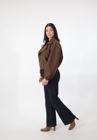 Brązowa krótka kurtka z oversize'owymi rękawami, dwiema guzikami, w zestawieniu z ciemnoniebieskimi jeansami o rozszerzanych nogawkach oraz brązowymi butami na wysokim obcasie. Jednolite białe tło.