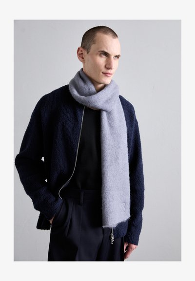 Bufanda de lana gris colgada alrededor del cuello, con una textura suave. Se lleva con una chaqueta azul marino texturizada y una blusa negra, con detalles de cremallera visibles.