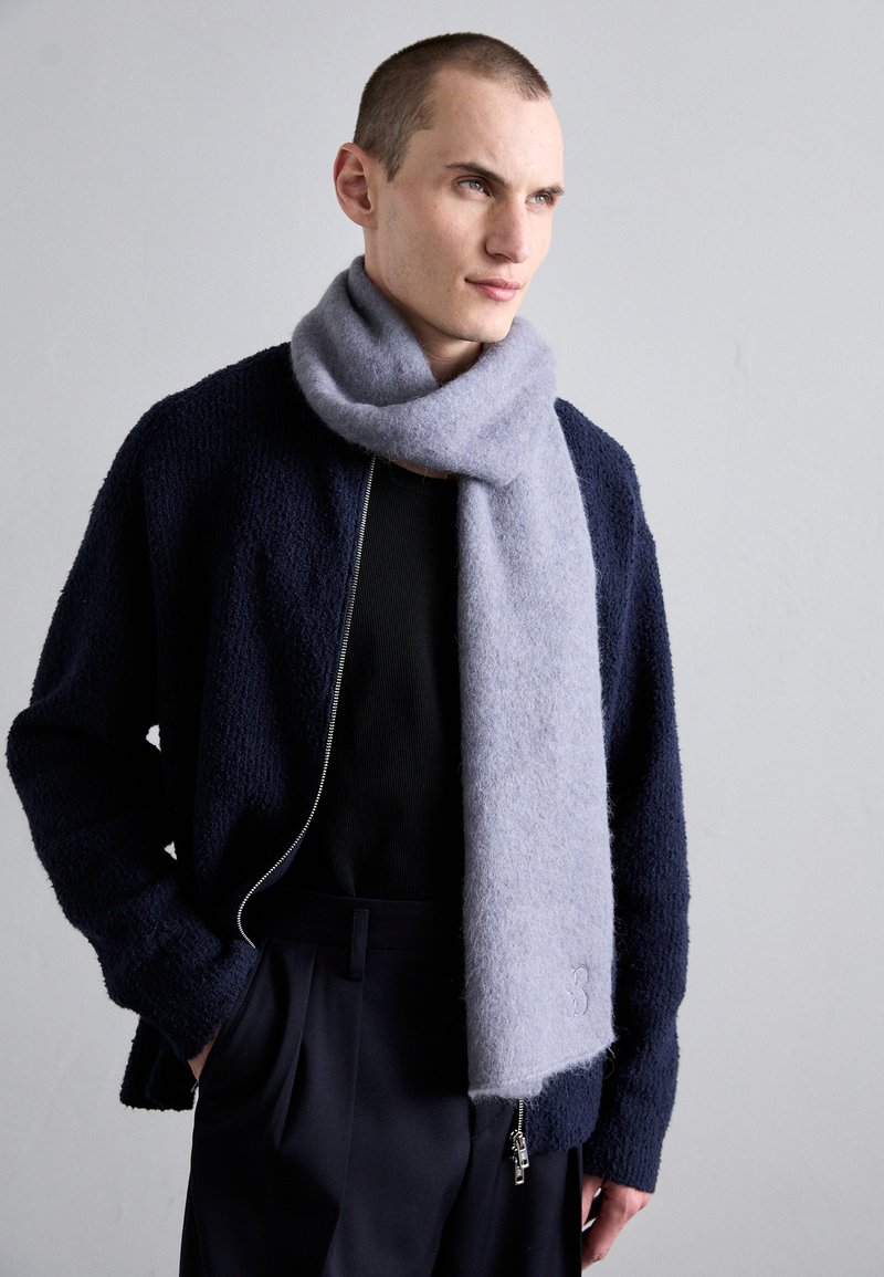 Pilka vilnos skara, nuvilkta aplink kaklą, pasižyminti minkšta tekstūra. Dėvima su tamsiai mėlynu tekstūriniu švarku ir juoda palaidine, matoma užtrauktuko detalė.