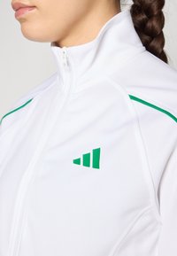 Valkoinen urheilullinen vetoketjullinen takki, jossa on vihreitä yksityiskohtia ja logo. Ominaisuuksiin kuuluu korkea kaulus ja sileä, kevyt kangas. Siisti muotoilu, ei kuvioita.