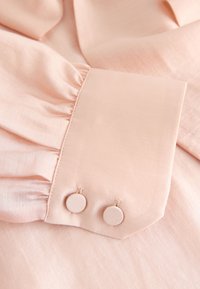 Gaiši rozā audums ar gludu tekstūru, kuram pievienots krošķelts aproce un divi apaļi pogas, pārklātas ar audumu, aizdarei.