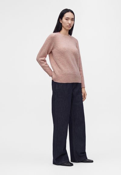 Femme portant un pull rose clair et un jean large foncé, debout avec une main derrière le dos sur un fond clair uni.