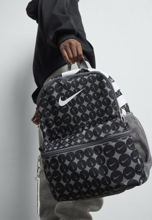 Oseba drži črno-sivo Nike nahrbtnik s krožnim vzorcem in belim logotipom swoosh pred enobarvno svetlo ozadjem.