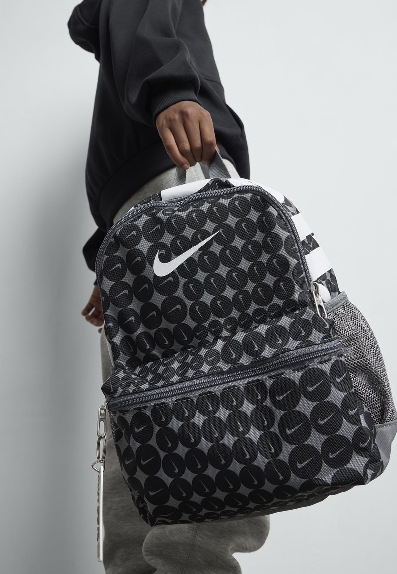 Άτομο που κρατάει μια μαύρη και γκρι σακούλα Nike με κυκλικό σχέδιο και λευκό λογότυπο Swoosh σε απλό φωτεινό φόντο.