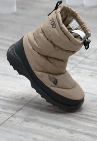 Botte isolante beige avec un design rembourré, semelle en caoutchouc et accents noirs. Présente un logo de marque et une fermeture réglable.