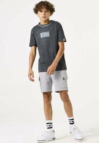 T-shirt grigia con stampa grafica, abbinata a shorts grigi chiari con tasche. Sneakers bianche e calze a righe completano l'outfit.