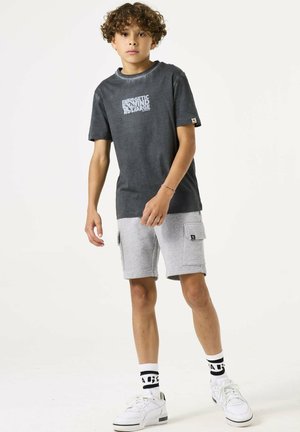 Graues T-Shirt mit grafischem Druck, kombiniert mit hellgrauen Shorts mit Taschen. Weiße Sneaker und gestreifte Socken vervollständigen das Outfit.