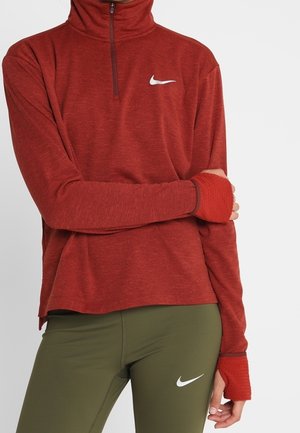 Osoba ubrana w czerwoną, dopasowaną bluzę Nike na suwak z długim rękawem oraz oliwkowozielone legginsy Nike, z otworami na kciuki w rękawach i widocznymi białymi logo Nike.