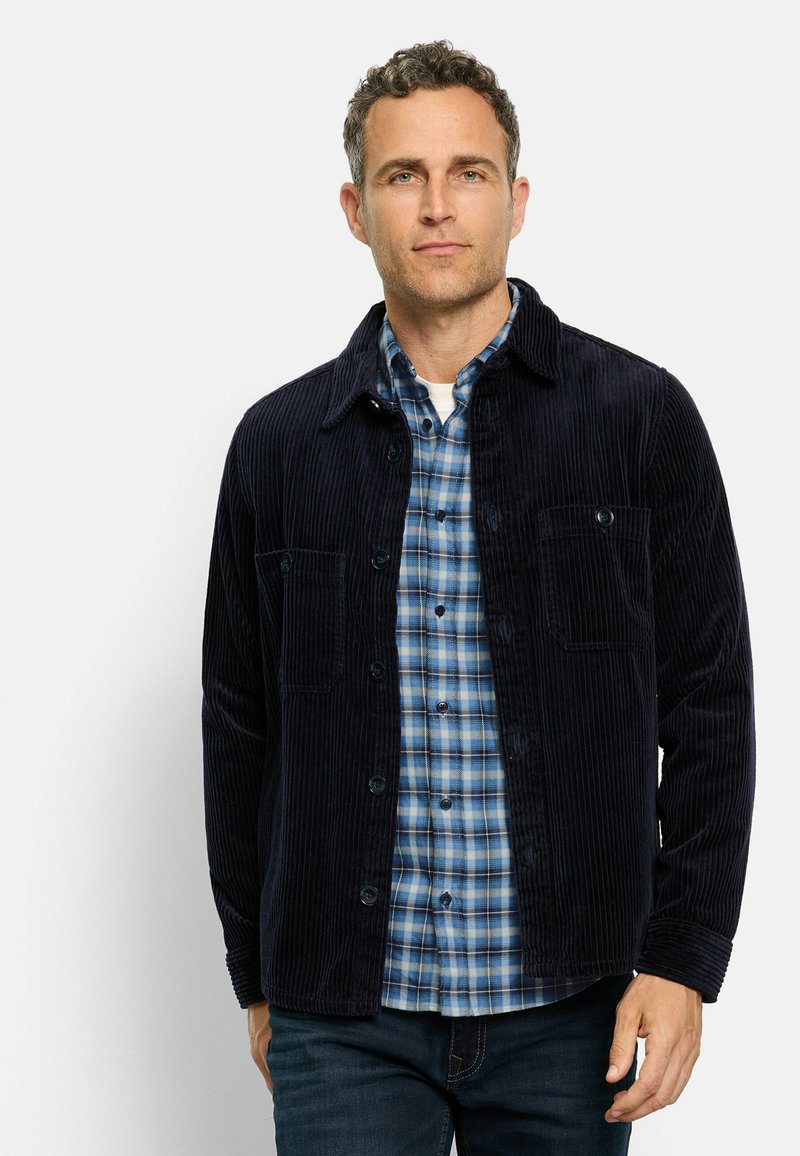 Marineblaue Cordjacke mit zwei Brusttaschen, Knopfverschluss und weicher Textur, kombiniert mit einem blau karierten Hemd.