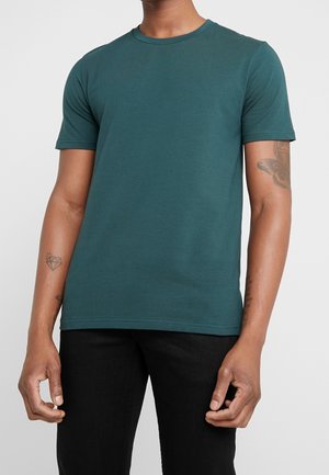Homme portant un t-shirt uni col rond à manches courtes de couleur bleu sarcelle et un pantalon noir, avec des tatouages visibles sur les deux avant-bras.