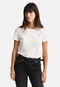 White cotton t-shirt with short sleeves featuring a small emblem on the chest, worn with high-waisted black jeans and a black belt.