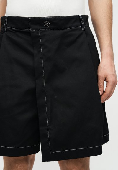 Μαύρα σορτς μέχρι το γόνατο με λευκή ραφή και μικρό μεταλλικό λογότυπο στη μέση, που φοριούνται από ένα άτομο με ορατό δεξί χέρι.