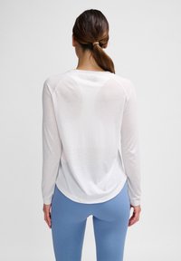 Vrouw met bruin haar in een paardenstaart, gekleed in een doorschijnend wit shirt met lange mouwen en blauwe legging, gezien van achteren tegen een effen achtergrond.