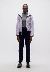 Γυναίκα με ανοιχτό μωβ μπουφάν, γκρι τοπ με φερμουάρ, σκούρο παντελόνι, μαύρα πεζοπορικά παπούτσια και μεγάλα αντανακλαστικά γυαλιά ηλίου, στέκεται μπροστά σε λευκό φόντο.