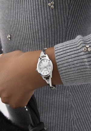 Silver watch with a round face and a textured white dial, featuring silver hour markers and a shiny metal bracelet. Worn on a hand holding a black bag.