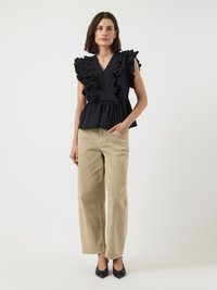 Donna che indossa una blusa nera senza maniche con rouches, pantaloni beige a gamba larga e tacchi neri a punta, in piedi contro uno sfondo bianco semplice.
