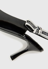 Black patent leather high-heeled shoe featuring a pointed toe and a logoed white elastic strap at the back. Thin, angular heel design.