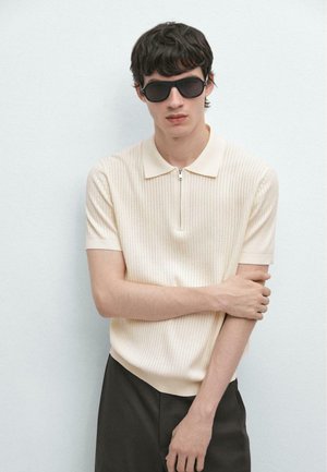 Giovane uomo con occhiali da sole neri, indossa una maglia a maniche corte color crema con zip e pantaloni neri, in piedi contro una parete chiara e semplice.