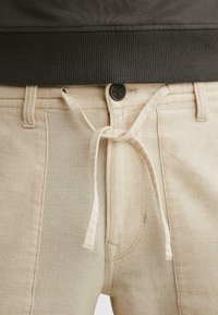 Beige gestructureerde broek met riemlussen, een zwarte knoop en een gebonden stoffen trekkoord, gedragen met een donkere geribde trui die ingestopt is.