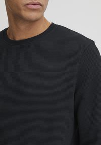 Schwarzer strukturierter Pullover mit rundem Ausschnitt, der ein dezentes gewebtes Muster aufweist. Der Stoff wirkt weich und leicht elastisch.